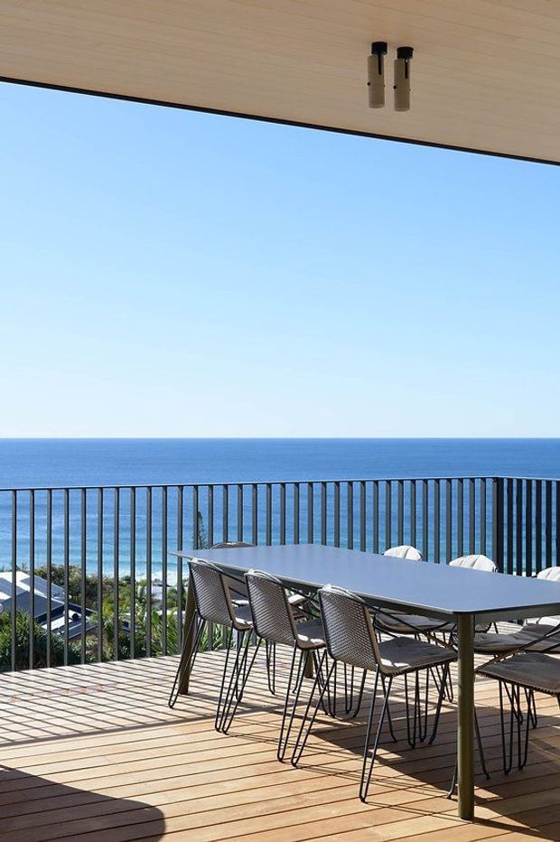A bright oceanfront terrace viewed from beneath a timber-lined ceiling. The spacious timber deck has a black metal balustrade overlooking the blue sea, with a long outdoor dining table and multiple chairs arranged for entertaining. Slim cylindrical ceiling lights and expansive open views contribute to a clean, contemporary coastal atmosphere.