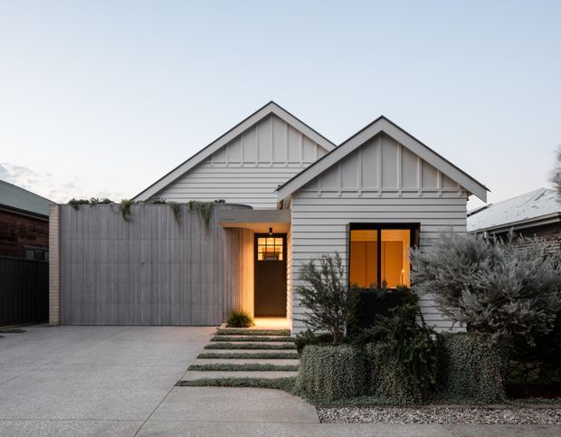 Exterior view of a compact residential home with two overlapping gabled volumes, white horizontal weatherboard cladding, decorative gable trim, dark-framed windows, and a warm-lit recessed entry. A concrete driveway and stepping-stone path lead through a low, drought-tolerant front garden, with a tall timber privacy fence screening part of the facade.