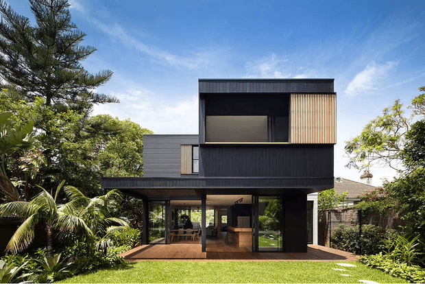A contemporary multi-level house set within lush tropical landscaping. The dark-clad upper storey features a projecting box form, a large recessed window, and a vertical timber privacy screen, while the lower level is largely glazed and opens onto a timber deck and lawn. Flat rooflines, strong geometric volumes, and sheltered outdoor living areas define the facade.