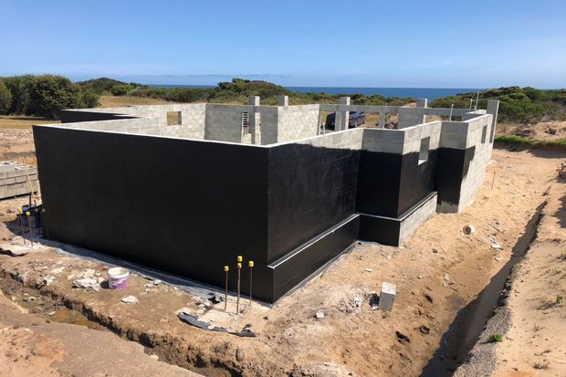 A low, partially constructed modern concrete-block house on a sandy coastal site. The exterior walls are being waterproofed or coated in black, while unfinished grey masonry walls and exposed service penetrations remain visible. Dense coastal vegetation and a blue ocean horizon appear beyond the building.