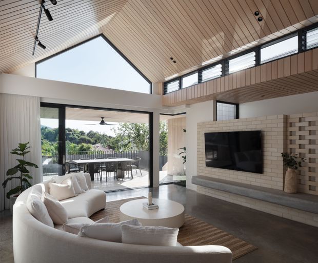 A bright, spacious contemporary living room with a curved cream sectional sofa, round coffee table, and wall-mounted television set within a pale brick feature wall. The room has a dramatic pitched timber-lined ceiling, clerestory windows, exposed upper-level void, and large glazed openings connecting to an outdoor dining terrace. Neutral finishes, indoor plants, and abundant natural light create an airy, refined atmosphere.