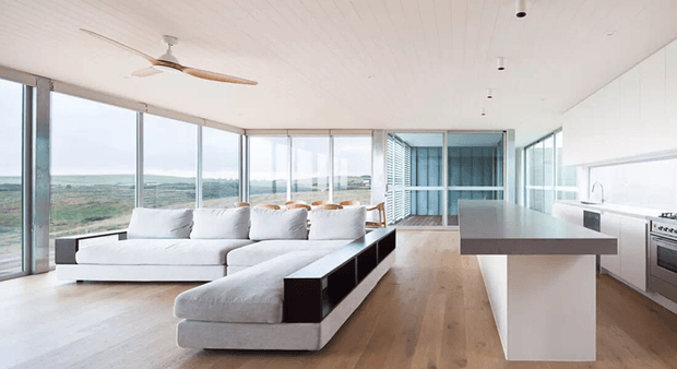 Open-plan contemporary living and kitchen space with expansive floor-to-ceiling glazing, a large light sectional sofa, pale timber flooring, white walls and ceiling, and a minimalist island kitchen. A broad landscape is visible through the windows.