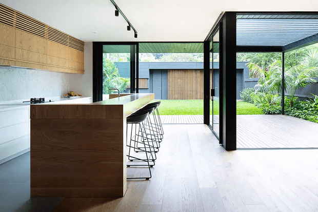A bright, open-plan contemporary kitchen with a large dark timber island and integrated bar seating, pale timber flooring, white cabinetry, and warm timber wall cabinetry. Black-framed floor-to-ceiling glazing opens directly onto a covered deck and landscaped courtyard, creating a strong indoor-outdoor connection. Linear track lighting and clean minimalist detailing reinforce the modern design.