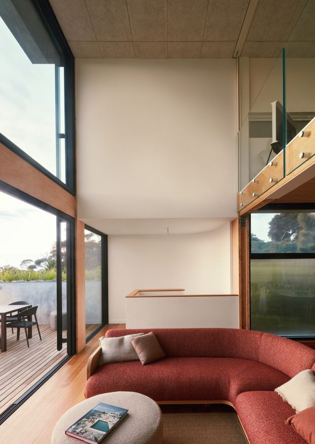 A warm, light-filled living room with a curved terracotta-red sectional sofa in the foreground, set against pale plaster walls and timber flooring. Full-height black-framed glazing opens onto a timber deck and landscaped exterior. A timber-and-glass balustrade at right overlooks the room, suggesting a mezzanine or upper-level landing. The ceiling has a subtle panelled finish, and the space has a refined contemporary character with natural materials and strong indoor–outdoor connection.