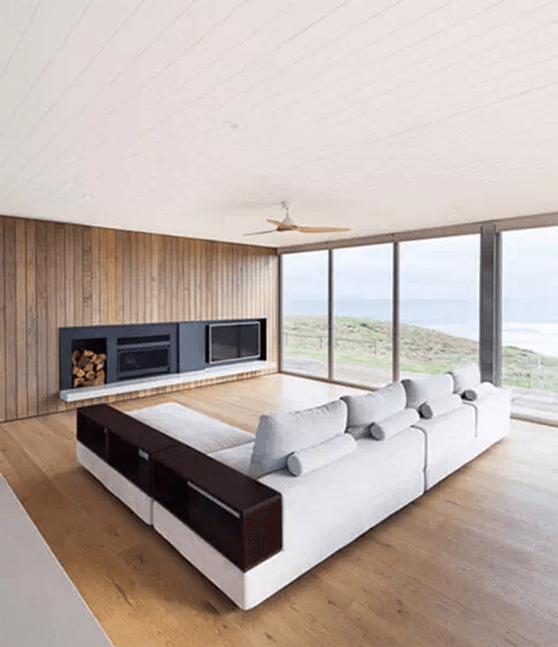 Bright contemporary living room with a large white sectional sofa, dark timber-backed built-in seating or console, a black fireplace and media unit set into a vertical timber feature wall, wide floor-to-ceiling glazing, timber flooring, and a low-profile ceiling fan. Coastal landscape is visible beyond the windows.