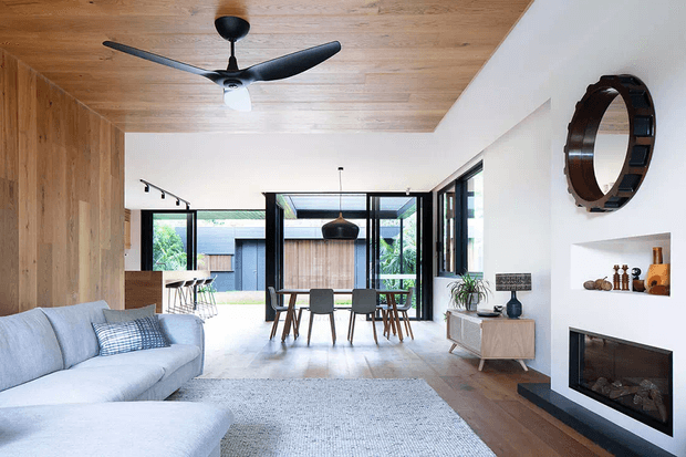 Open-plan contemporary living and dining area with warm timber wall and ceiling lining, light wood flooring, a pale sectional sofa, dining table and chairs, black-framed glazing, and a built-in fireplace set into a white wall. The room opens visually toward a landscaped courtyard and features a sculptural ceiling fan and circular wall clock.