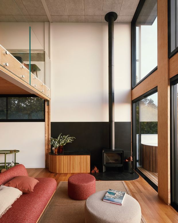 A warm, double-height living room with a loft/mezzanine visible at left, extensive black-framed glazing, timber structural framing and paneling, and a freestanding black wood-burning stove. The room features a rust-red sofa and ottoman, a textured cream coffee table, timber flooring, a ribbed timber console, and a neutral rug. The space has a strong indoor-outdoor connection and abundant natural light.