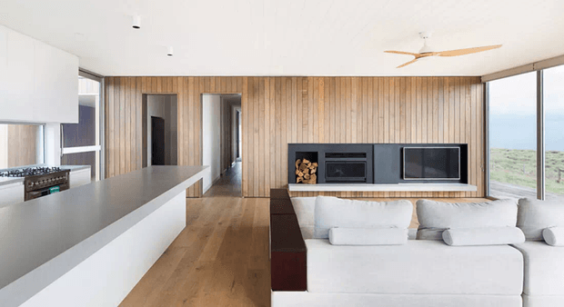Open-plan contemporary interior combining a minimalist white kitchen with a long island, warm vertical timber wall panelling, timber flooring, and a white sectional sofa facing a built-in media and fireplace wall. Full-height glazing on the right provides expansive landscape views, while a ceiling fan and recessed lighting complete the clean, light-filled space.