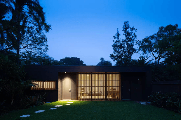 A low, single-storey modern residence photographed at dusk, with a dark flat-roofed facade, warm illuminated floor-to-ceiling screened glazing, a recessed entry, and a manicured lawn with stepping stones. Mature vegetation surrounds the building, creating a tranquil garden setting.