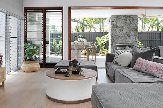 Bright, contemporary living room opening onto an outdoor patio or courtyard through wide glazed doors. The room features pale timber flooring, white horizontal wall cladding, dark timber-framed louvred glazing, a large grey sectional sofa, and a round white coffee table with a timber base. A stacked natural-stone fireplace forms a prominent feature wall, while outdoor dining and planting are visible beyond.