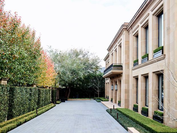 Formal landscaped driveway or courtyard beside a large symmetrical stone-clad building. The facade features tall recessed windows with strong classical proportions, decorative mouldings, projecting cornices, window boxes, and a small balcony. Neatly clipped hedges, espaliered greenery, mature trees, and pale grey paving create a refined institutional or luxury residential approach.