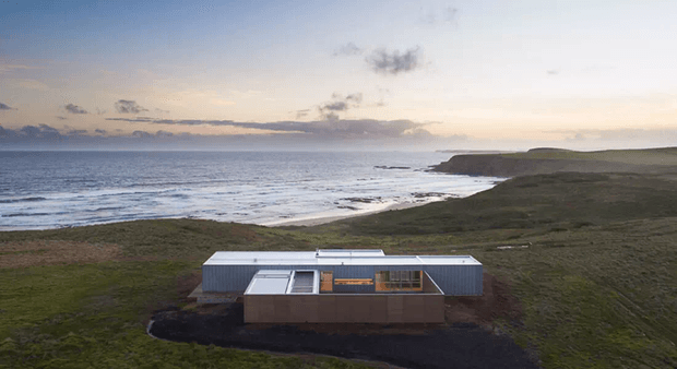 A low, contemporary coastal house set in an open grassy landscape above the ocean. The building has a compact rectangular form, extensive glazing, a flat or very low-pitched roof, and pale corrugated metal-clad side volumes with a darker central facade. Waves, a rugged shoreline, and a broad ocean horizon dominate the setting.