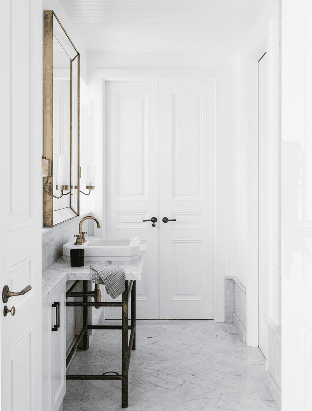 Bright, narrow bathroom or powder-room interior viewed from the doorway, with white paneled walls and double doors, a marble-look tiled floor, and a compact white vessel sink on a dark metal-framed console. A large aged-metal framed mirror, wall sconces, exposed plumbing, and a grey stone or marble benchtop create a refined classic look.