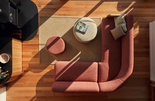 Top-down view of a warm, sunlit living room with a large rust-red upholstered sectional sofa arranged around a textured beige area rug. A round cream ottoman and smaller rust-red stool sit centrally, with dark built-in cabinetry and a ribbed timber-fronted unit along the left edge. The room has honey-toned timber flooring and ceiling, strong diagonal sunlight and shadows, and a cozy contemporary feel.