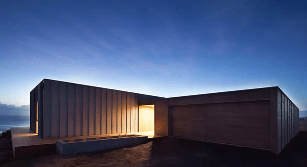 A low, angular modern building viewed externally at dusk. The composition features dark vertically ribbed metal cladding, a large recessed timber-toned garage or entry door, a warm illuminated recessed entrance, flat rooflines, and a minimal paved or gravel forecourt against a blue evening sky.