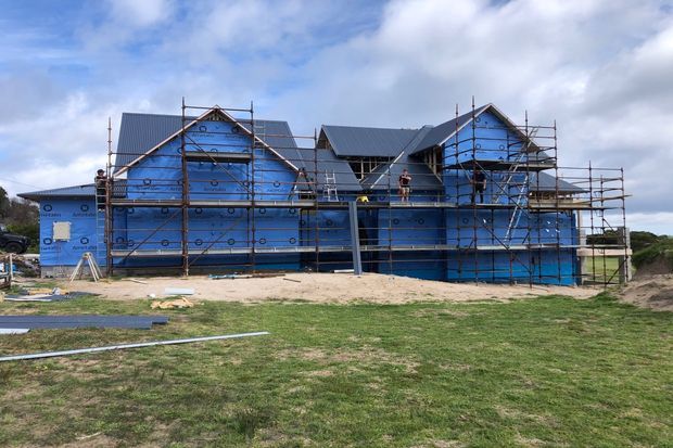 Exterior view of a large multi-gabled house under construction, wrapped in blue building membrane and surrounded by extensive scaffolding. Dark pitched metal roofs and partially installed exterior elements are visible, with an open grassy construction site in the foreground.