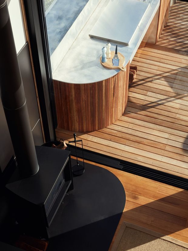 A close architectural detail showing a dark freestanding wood-burning stove beside full-height glazing, opening onto a timber deck. Outside is a rounded vertical timber-clad structure with a pale curved roof or benchtop, while warm timber flooring continues indoors. Strong sunlight creates geometric shadows across the deck and interior.