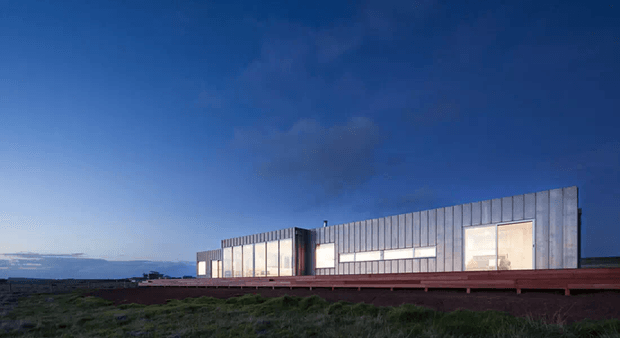 A long, low modern building viewed from an open grassy landscape at dusk. The facade features extensive vertical metal cladding, large illuminated rectangular windows, and a dark red horizontal base or terrace element beneath a flat roofline. The composition emphasizes the minimalist linear architecture against a deep blue sky.