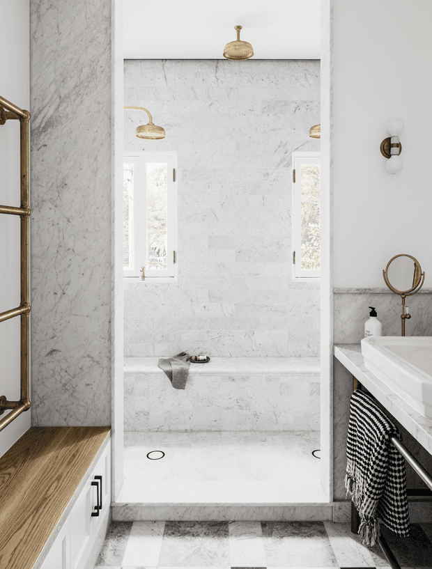 Bright, refined bathroom interior centered on a marble-lined walk-in shower with twin windows and brass shower fittings. A white vanity with marble splashback and countertop occupies the right side, while white cabinetry with a timber bench runs along the left. Brass sconces, a round shaving mirror, monochrome textiles, and a patterned marble floor complete the space.