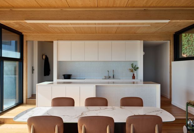 A warm, compact contemporary kitchen and dining area with extensive light timber lining to the ceiling and beams, white flat-panel cabinetry, a pale textured tiled splashback, and a large light stone island with integrated cabinetry. A marble-look dining table with brown upholstered chairs occupies the foreground. Dark-framed glazing on both sides brings in natural light, with timber flooring and minimal decor.