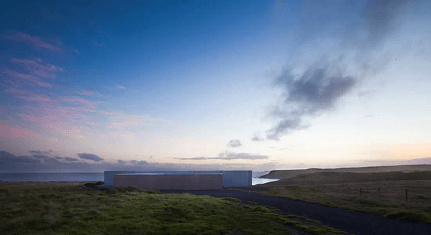 A low, minimalist rectangular building sits in a rugged coastal landscape at dusk, with dark paving leading toward it and the sea visible beyond. The simple box-like form has a muted grey-blue exterior and is set against open grassy terrain, dramatic clouds, and a softly illuminated horizon.