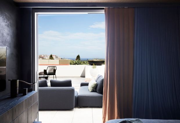 A contemporary interior view looking through a wide glazed opening toward a bright outdoor terrace. The foreground is a dark, moody living or media area with a large television, dark timber-toned built-in cabinetry, textured dark wall surfaces, and a full-height curtain. Beyond it is a light-filled outdoor lounge with grey sofas, a chair, pale tiled flooring, and a low white parapet framing a distant coastal or urban view.