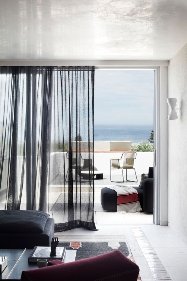 A bright contemporary living room opens directly onto a tiled terrace with expansive ocean views. Sheer black curtains frame large sliding glass doors, while dark sofas, a burgundy armchair, a black coffee table, and a patterned rug create contrast against the white textured walls, white ceiling, and pale flooring. A sculptural wall light and minimalist outdoor seating are visible.