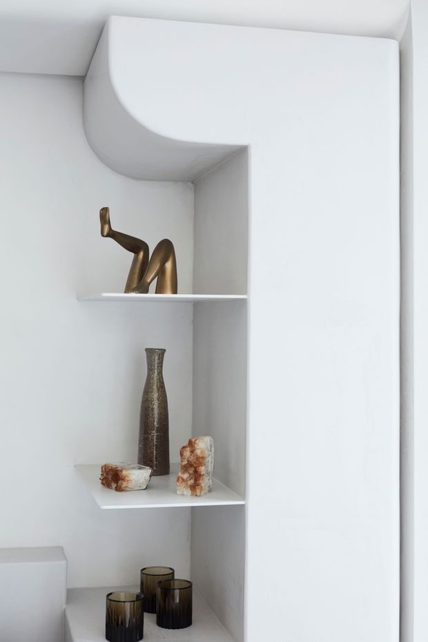 Close-up of a minimalist white built-in recessed shelving unit with softly curved upper detailing and integrated open shelves. The shelves display a bronze-toned sculptural object, a tall dark textured vase, pale stone-like decorative objects, and dark glass vessels. A partial white sanitary fixture is visible at lower left, suggesting the shelving is in a bathroom. The overall composition is clean, monochromatic, and sculptural.