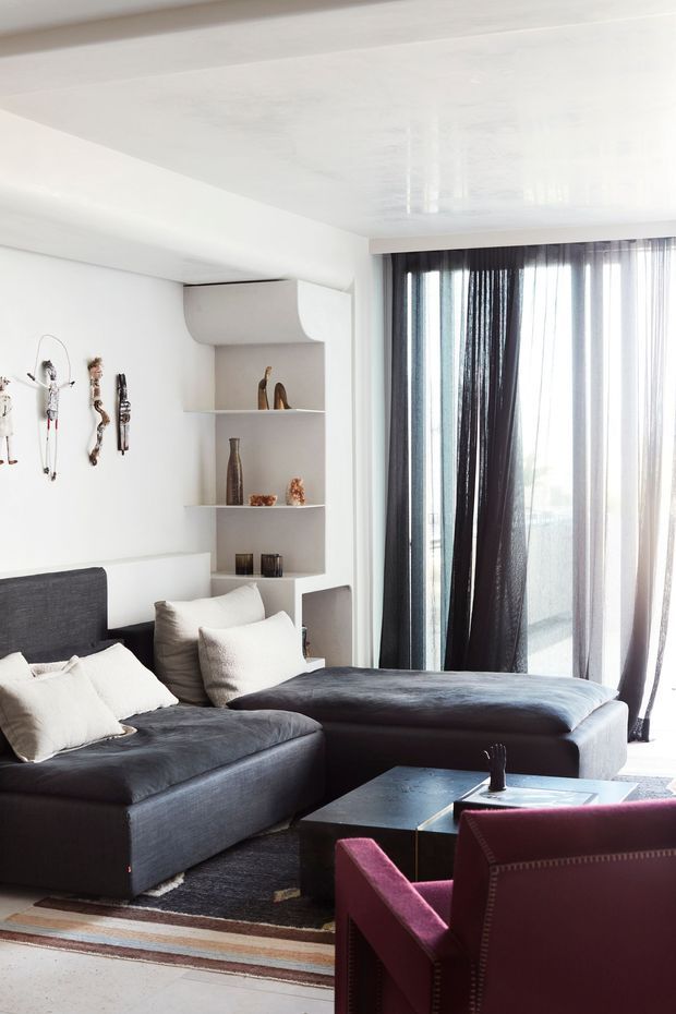 A bright contemporary living room with a large dark charcoal sectional sofa, white textured cushions, a black coffee table, and a burgundy accent armchair. Full-height sheer and dark curtains cover a glazed opening, while a recessed built-in shelving unit displays decorative objects. The room has white walls and ceiling, layered area rugs, and a clean, refined monochromatic palette with deep accent colours.