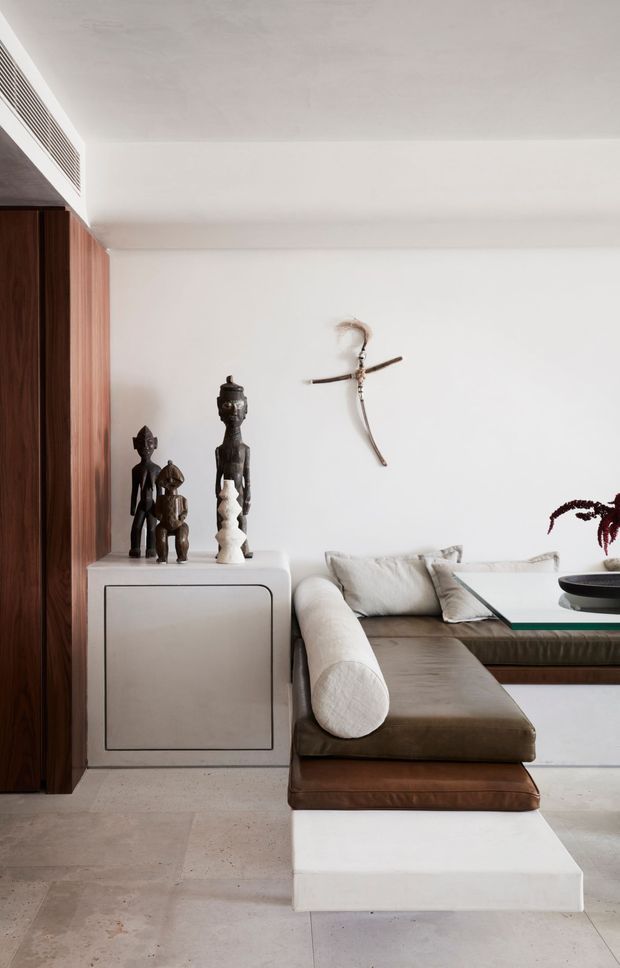 A refined contemporary living room with a low modular sofa in taupe and brown leather, positioned beside a white built-in cabinet and a dark timber wall panel. Sculptural tribal-style objects sit on the cabinet, while a minimalist wall-mounted decorative piece and a low glass-topped coffee table complete the space. Large pale stone or concrete-look floor tiles and white plaster walls create a calm, warm-neutral interior.