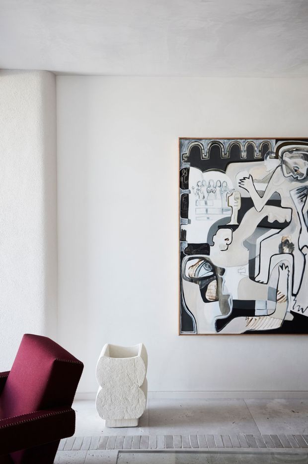A refined, minimalist interior corner with white textured walls and ceiling, a pale stone or concrete floor, and a large monochrome figurative artwork in a simple frame. A sculptural white vessel stands below the artwork, accompanied by a burgundy upholstered armchair with visible nailhead trim. The room has a quiet gallery-like atmosphere and restrained contemporary styling.