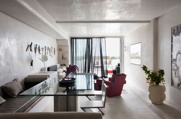 Bright open-plan contemporary living and dining area with a refined neutral palette. The room features a glossy reflective ceiling, pale textured walls, large light-coloured floor tiles, a glass dining table, built-in cabinetry, a low sofa, sculptural coffee tables, and two burgundy lounge chairs. Sheer black curtains frame full-height glazing and a balcony beyond. Artwork, wall-mounted decorative objects, and indoor plants add character.