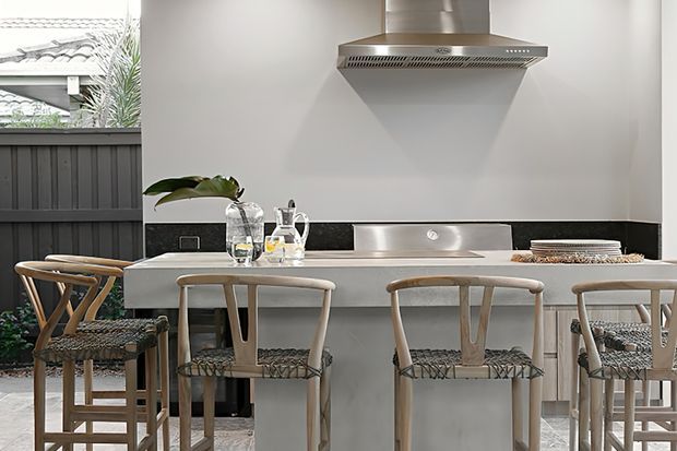 Bright contemporary kitchen with a central island and integrated stainless-steel cooktop, rangehood and splashback. Light timber bar stools with woven seats line the island, while dark cabinetry or fencing and greenery are visible beyond the open side of the space.