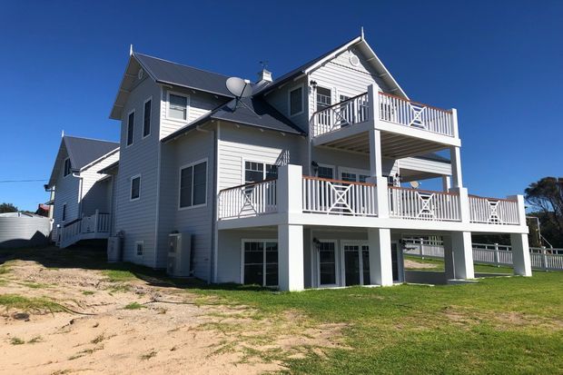 A large multi-level coastal-style house with white horizontal weatherboard cladding, dark pitched gabled roofs, and several stacked verandas or balconies featuring white balustrades with cross-braced detailing. The home sits on a gently sloping sandy and grassy site under a clear blue sky.