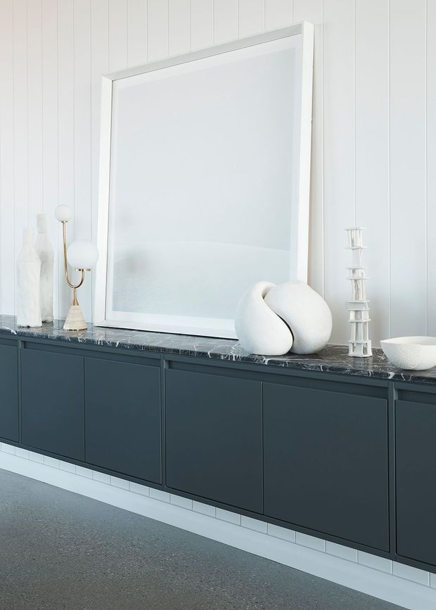 A bright, highly styled interior vignette featuring a long wall-mounted dark blue-grey storage cabinet with a polished dark marble benchtop. White vertical wall panelling, a large white-framed mirror or artwork, and sculptural white decorative objects create a crisp, refined monochromatic composition. A pale terrazzo-like floor and white tiled plinth complete the scene.