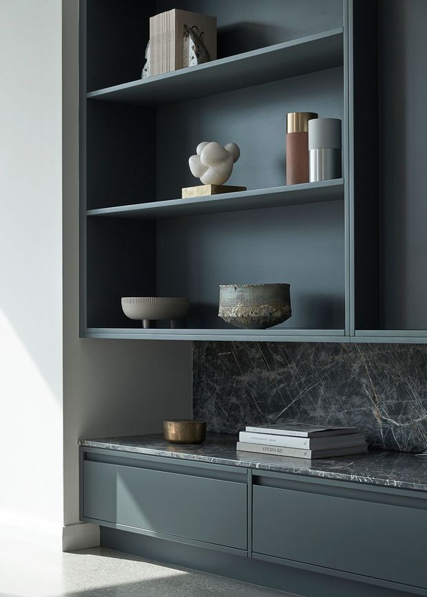 Close-up interior view of a sophisticated dark grey built-in cabinetry and open shelving unit. The joinery includes two display shelves with decorative objects, lower drawers, a dark veined stone benchtop, and a matching stone splashback. Natural light creates strong shadows across the muted, refined composition.