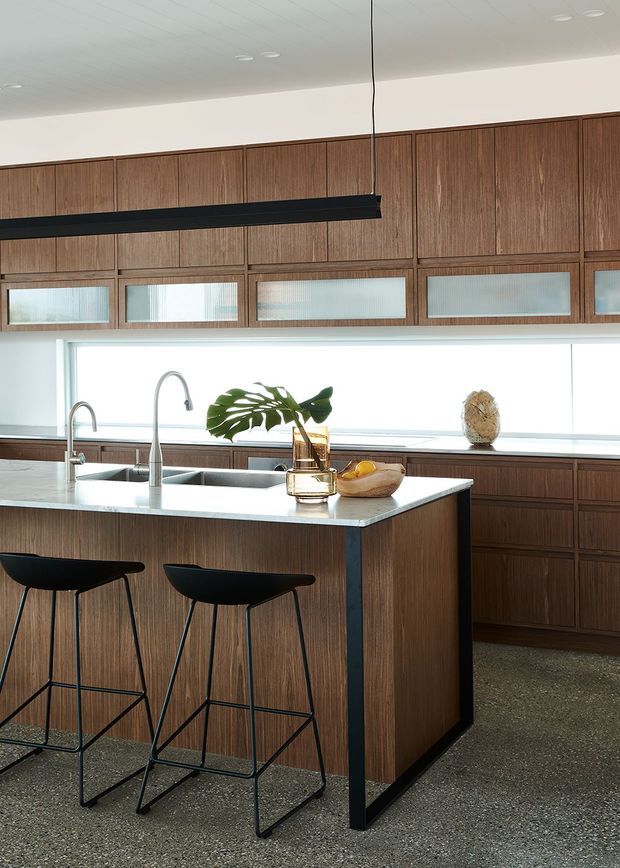 Contemporary kitchen with extensive warm timber cabinetry, a large pale stone island with integrated double sink and gooseneck tap, and two black bar stools. A long black linear pendant hangs above the island, while high clerestory-style glazing and a bright window provide abundant natural light. The floor appears to be dark grey polished concrete or terrazzo.