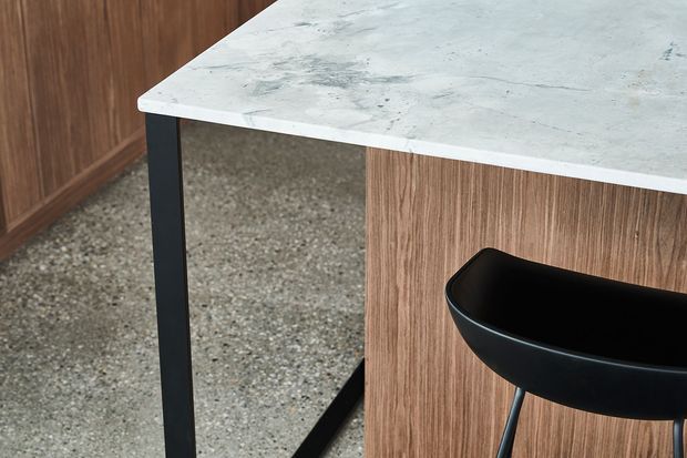 Close-up of a contemporary kitchen island or bar counter featuring a pale veined stone benchtop, vertical timber joinery, a black metal support frame, and a black bar stool. A speckled hard-surface floor and timber cabinetry are visible in the background.