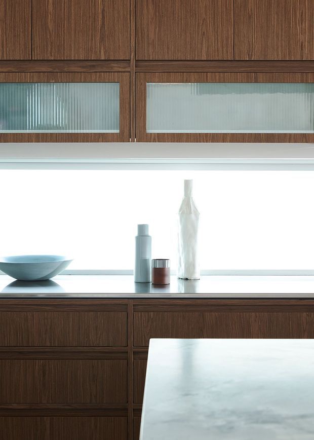 Close-up of a refined kitchen interior featuring dark walnut-toned timber cabinetry with integrated drawers and upper cupboards, ribbed translucent glass cabinet fronts, a pale stone benchtop, and a bright frosted window or splashback area. Minimal decorative objects sit on the counter, with a light stone island or work surface in the foreground.