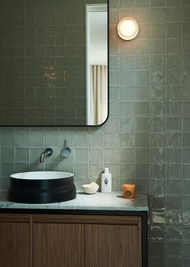 A refined bathroom vanity featuring a large round black vessel sink on a pale stone benchtop, dark timber cabinetry, brushed metal wall-mounted tapware, and glossy green-grey square wall tiles. A rounded-corner mirror and small wall light sit above the vanity, with minimal toiletries adding warmth and texture.