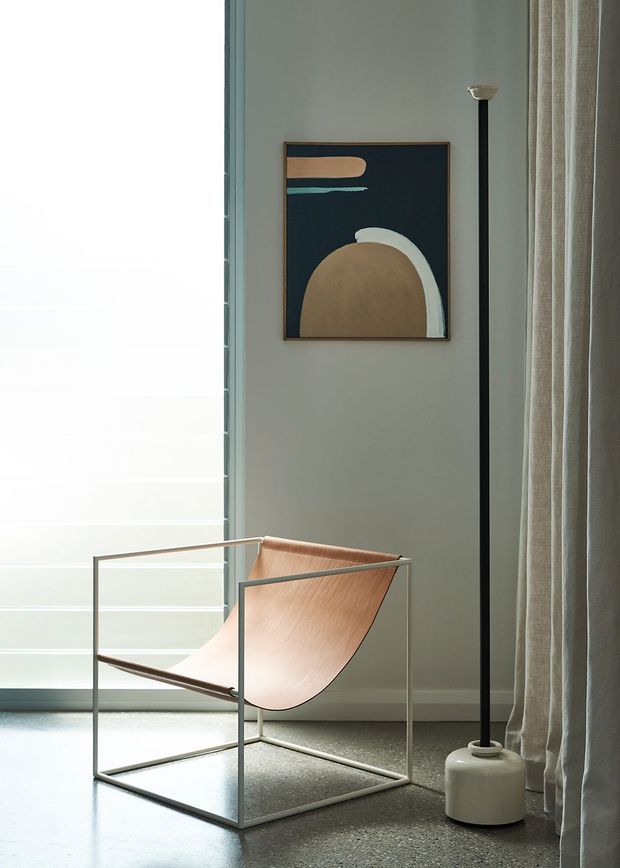 A softly lit, minimalist interior vignette featuring a sculptural tan leather sling chair with a slim metal frame, positioned beside a tall black floor lamp. A framed abstract artwork hangs on a muted grey wall, with sheer floor-to-ceiling curtains and a large frosted or translucent window creating diffused natural light. The floor has a speckled polished concrete or terrazzo-like appearance.