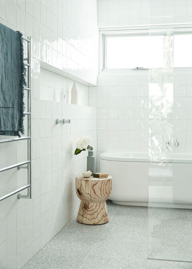 Bright contemporary bathroom featuring white glossy square wall tiles, a freestanding white bathtub behind a clear glass shower screen, a chrome heated towel rail with a dark towel, and a pale grey terrazzo-style floor. A sculptural natural timber stool adds warmth beside the bath. Large high-level glazing provides abundant natural light.