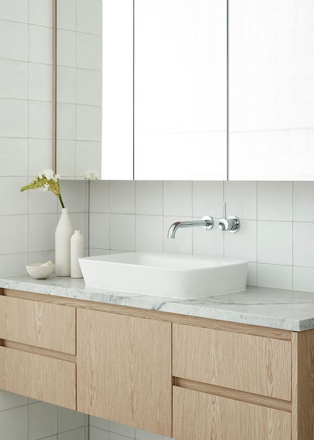 A bright, minimalist bathroom vanity featuring a wall-mounted light timber vanity with integrated drawers, a white vessel basin, wall-mounted chrome tapware, pale tiled walls, and a pale veined stone benchtop. A large mirrored cabinet occupies the upper wall, with simple white ceramic vases as restrained decor.