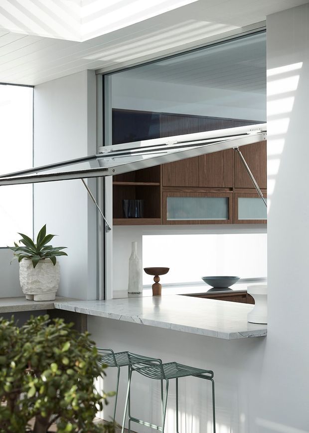 Bright contemporary kitchen servery with a raised glass awning window opening to a counter, white marble-look benchtop, dark timber overhead cabinetry with ribbed glass panels, and minimalist metal bar stools. Strong daylight enters through the large glazed opening, with a potted plant and restrained decorative objects adding a natural accent.