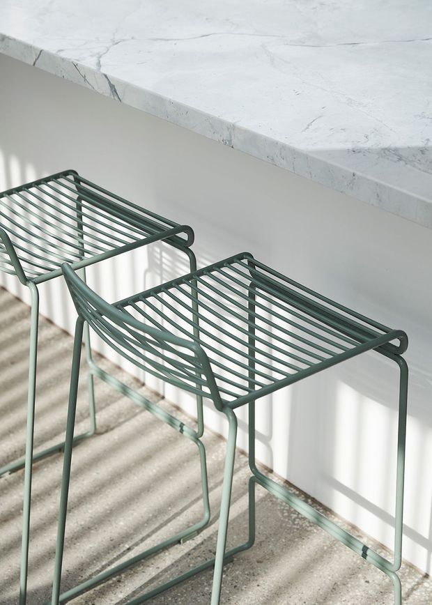 Close-up interior detail of two sage-green metal wire bar stools positioned beside a pale stone countertop or island. Strong sunlight creates striped shadows across the light wall and terrazzo-like floor, giving the scene a clean, airy contemporary feel.