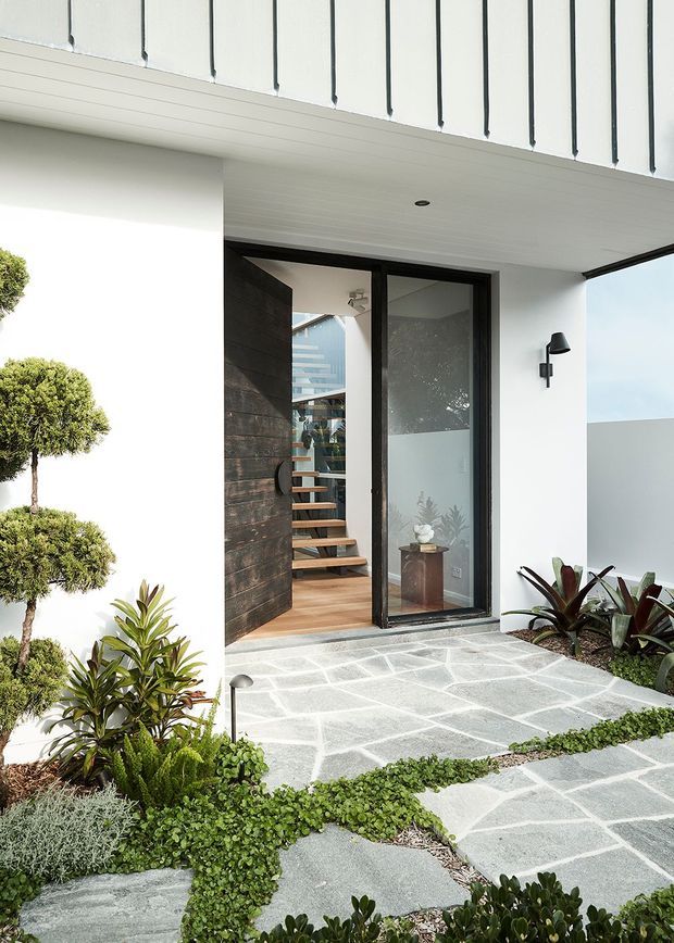 Contemporary residential entry viewed from an outdoor landscaped approach. The facade is crisp white with black vertical detailing and dark-framed glazing. A large timber pivot door opens to reveal a warm timber-floored stair hall with floating stairs. The entry path is paved in irregular pale grey stone slabs and surrounded by sculptural clipped greenery and low planting.
