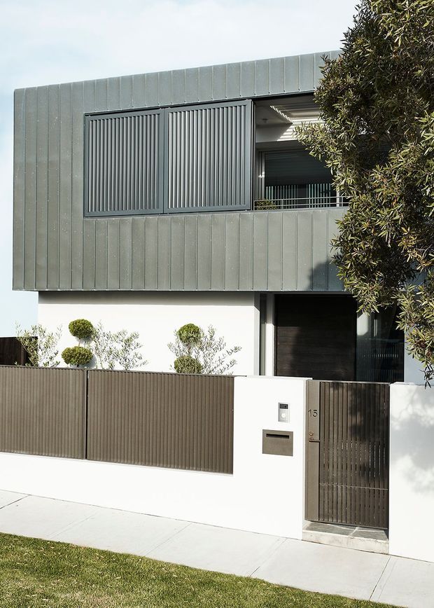 Street-facing view of a contemporary multi-storey residence with a clean rectilinear facade. The upper level is clad in vertically seamed dark grey metal panels with matching vertical privacy screens and balcony balustrade. The lower level is white rendered masonry, with a dark timber or metal entry door, gated pedestrian entry, and a simple front boundary fence. Trimmed topiary and a street verge appear in the foreground.