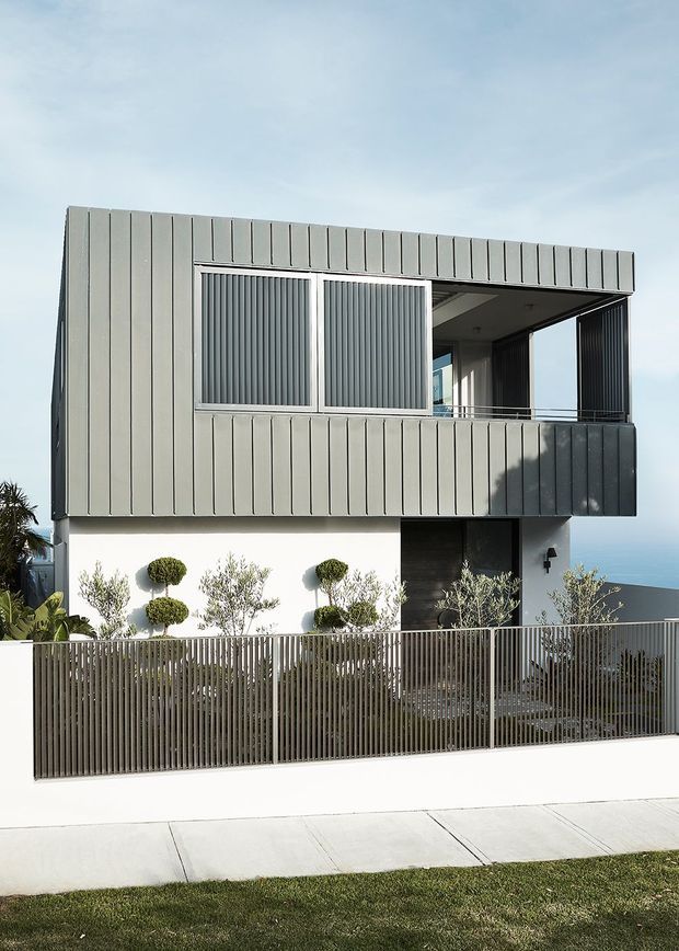 A contemporary two-storey residential home with a box-like form, clad in vertically ribbed grey metal panels. The upper level features sliding louvred screens, a recessed balcony, and dark-framed glazing, while the lower level has white rendered walls, dark cladding around the entry, and a landscaped front garden behind a slim vertical metal fence. The property sits beside a paved footpath, with a glimpse of blue water visible beyond the building.