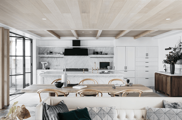 A bright open-plan kitchen, dining and living area with a predominantly white and pale timber palette. The kitchen features a marble-look backsplash and island, white built-in cabinetry, black appliances and brass-toned fixtures. Pale timber ceiling boards and exposed beams add warmth, while a large timber dining table with curved timber chairs sits behind a tufted white sofa. Tall black-framed glazing is visible along the left side.