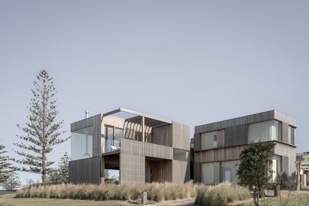 Exterior photograph of a modern multi-storey residential building composed of offset rectangular volumes. The facade combines dark vertical cladding, lighter textured panels, extensive glazing, recessed balconies, and flat rooflines. Ornamental grasses and mature trees surround the building, with a pale overcast sky in the background.